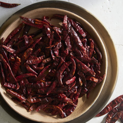 A wooden bowl filled with numerous dried Chile de Arbol chilies sits on a light-colored, textured surface. Several fiery Mexican chiles have spilled out onto the table, their vibrant hues and shapes highlighted by soft shadows.