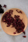 A round wooden bowl holds a loose pile of dried red Hibiscus Flower petals, known as Flor de Jamaica, some of which spill onto a light gray stone surface. The crinkled petals are deep red with slight yellow tips, and a few lie outside the bowl.