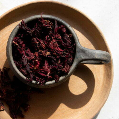 A rustic ceramic cup filled with dried Hibiscus petals sits on a round, tan clay plate. Some Flor de Jamaica petals spill onto the plate, while natural sunlight casts soft shadows on the light, textured surface.