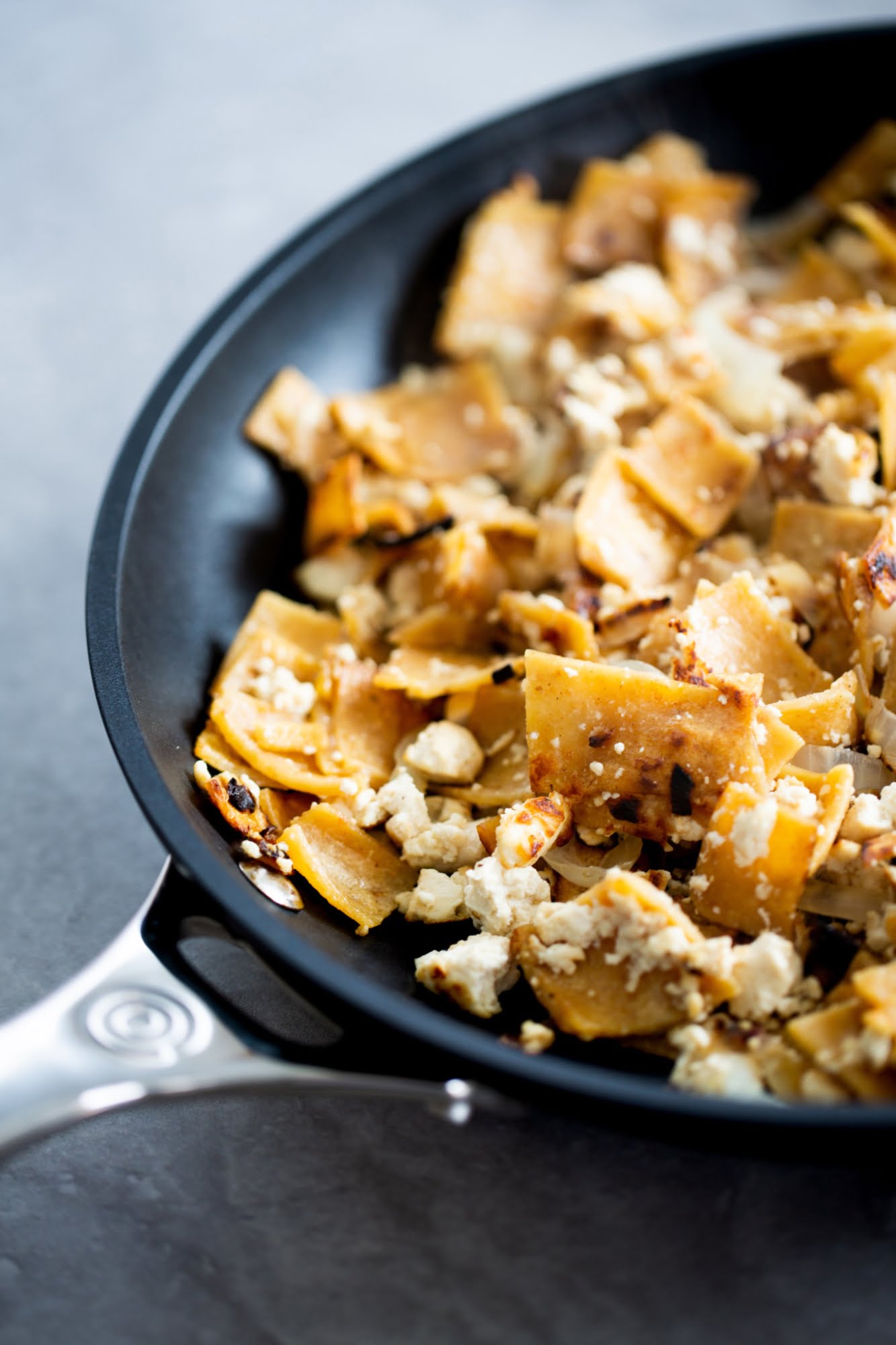 A close-up of a black frying pan with golden, crispy tortilla pieces and crumbled white cheese on top. The pan sits on a gray surface, and the food appears freshly cooked, with some browned edges on the tortillas.