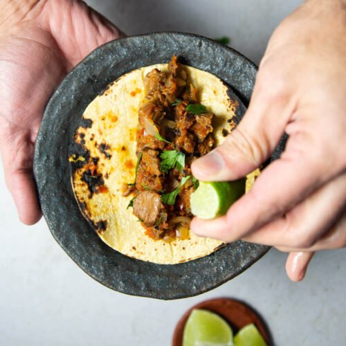 A hand squeezes a lime wedge over plant-based Mexican tacos filled with seasoned meat and herbs on a small, rustic plate. The other hand holds the plate. Below, a dish with lime wedges sits on a light-colored surface.