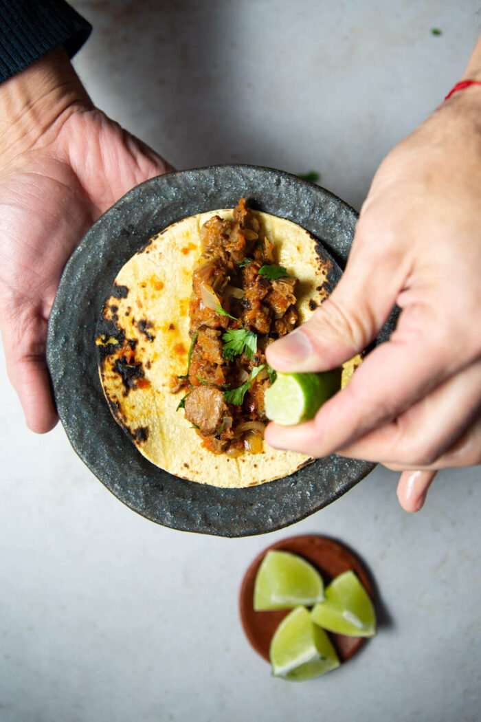 Man holding dark dish with bistec a la mexicana over a corn tortilla and some lime being squeezed