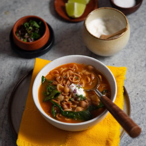 A bowl of soup with noodles, chickpeas, greens, and a dollop of cream—reminiscent of comforting sopa de conchas—sits on a plate with a yellow napkin. Nearby are lime wedges, salt, fresh herbs, and sour cream.
