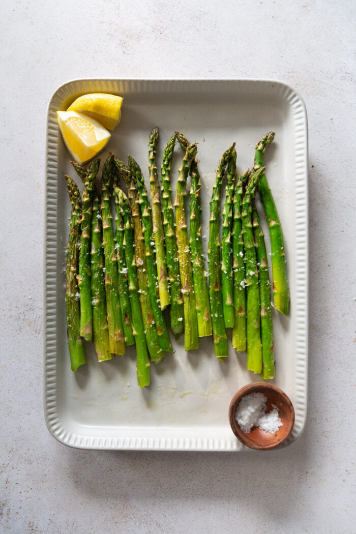 Asparagus on a gray platter on top with a bowl of salt and a lemon.