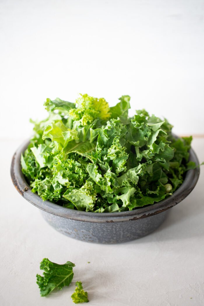 A rustic gray bowl filled with fresh, curly green kale leaves sits on a light surface. A few stray kale pieces rest beside the bowl. The background is white and minimal, drawing focus to the vibrant, textured leaves.