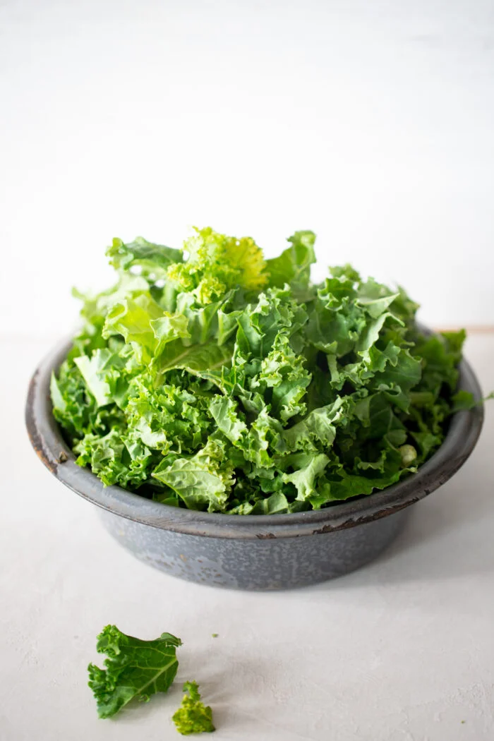 A rustic gray bowl filled with fresh, curly green kale leaves sits on a light surface. One piece of leafy kale has fallen beside the bowl&mdash;perfect for adding to your favorite recipe, like salads or vibrant chia overnight oats.