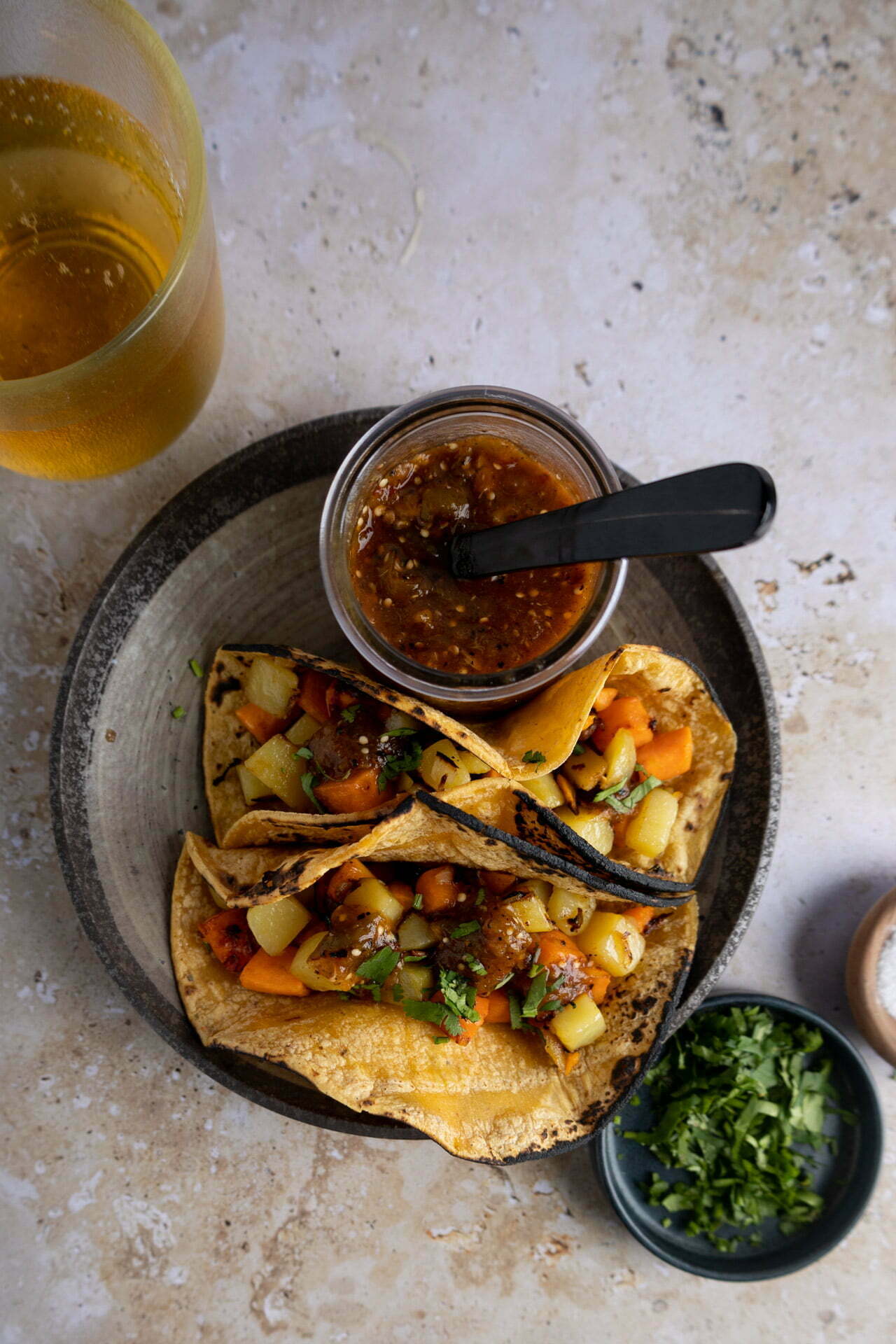 A plate with two vibrant Vegan Tacos filled with colorful diced vegetables sits beside a small jar of salsa. A glass of yellow drink, a bowl of chopped herbs, and a wooden dish of salt rest nearby on the beige surface.