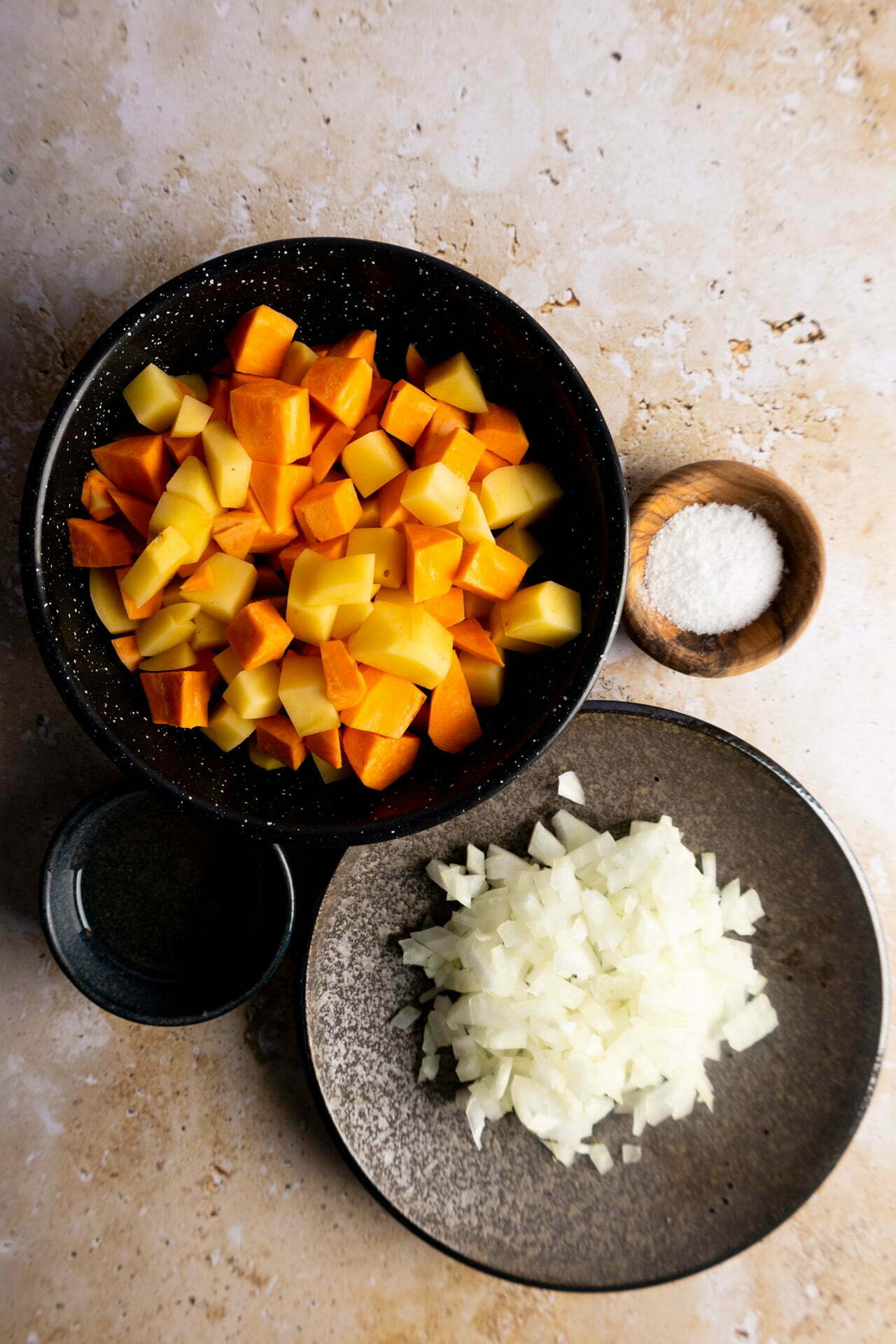 A flat lay of diced sweet potatoes and potatoes in a black bowl hints at delicious Vegan Tacos, with chopped white onions, a small wooden bowl of salt, and a cup with liquid arranged neatly on a light, speckled surface.
