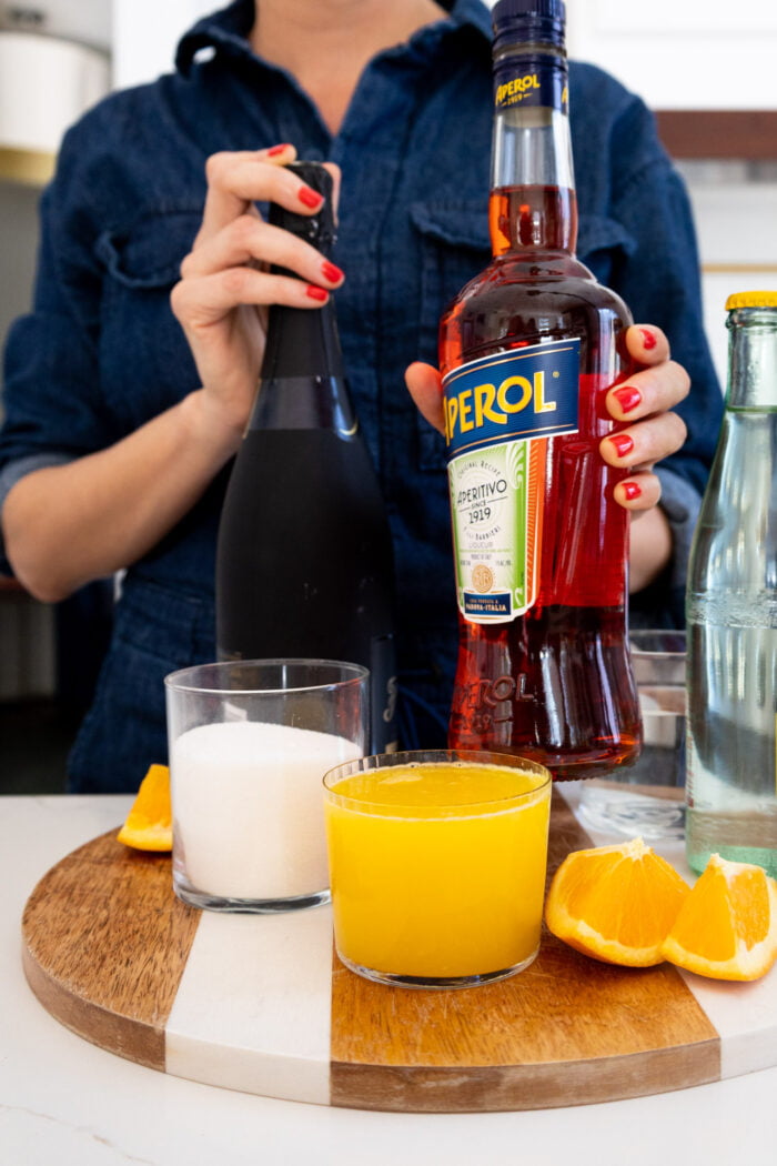 Una botella de aperol, proseco, agua con gas y un vaso de zumo de naranja