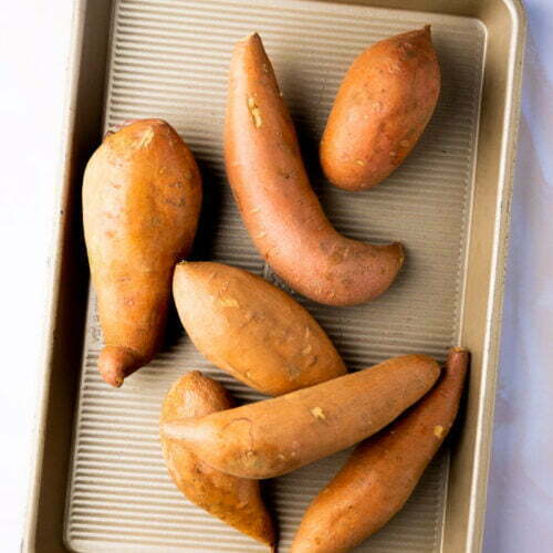 A baking sheet holds seven whole, unpeeled sweet potatoes of varying sizes and shapes, perfect for baked sweet potatoes or trying new sweet potato recipes. The metallic, ridged sheet sits on a light-colored background.