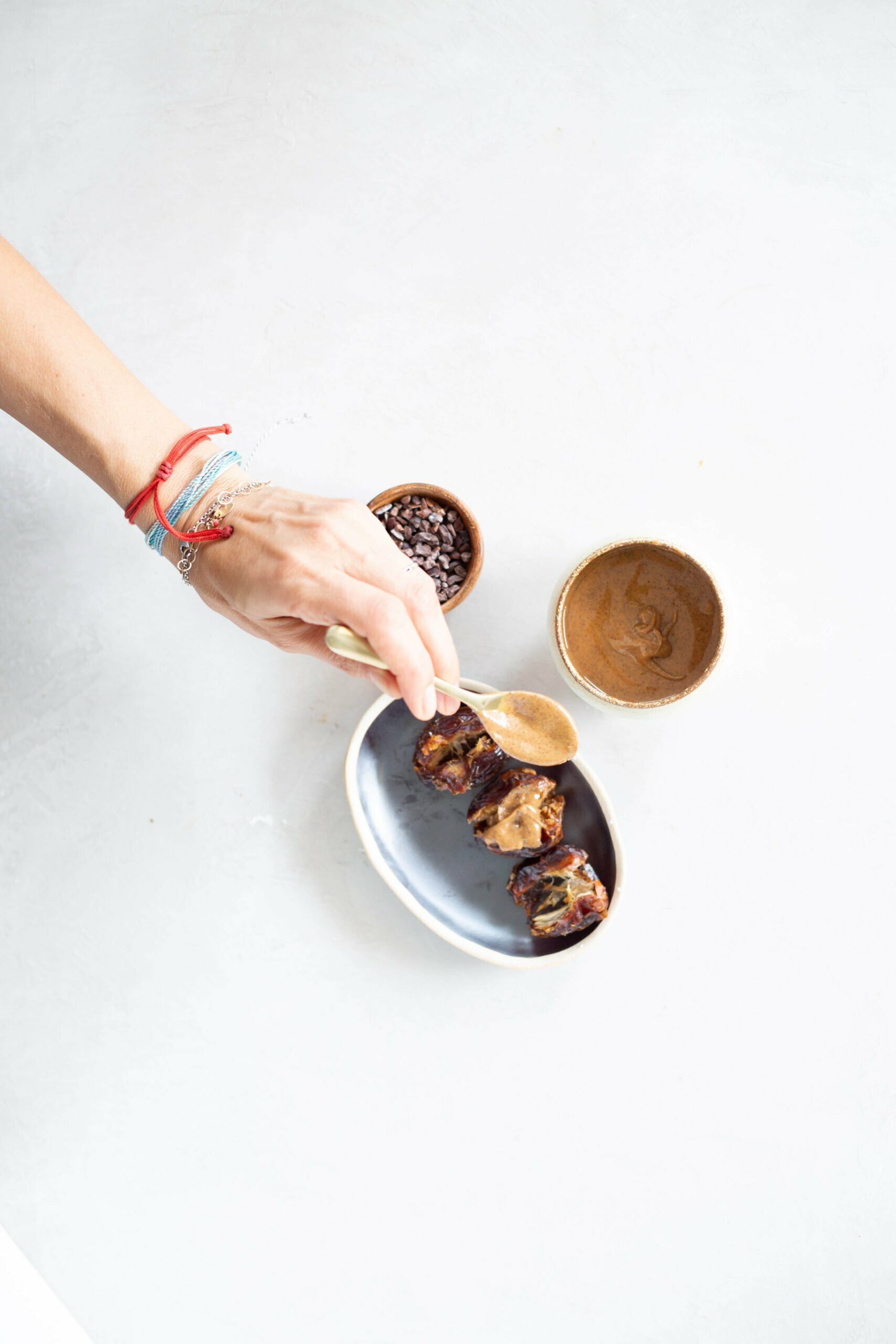A hand with colorful bracelets holds a gold spoon, scooping almond butter onto stuffed dates in a small bowl. Nearby are two small dishes: one with cocoa nibs and one with smooth nut butter, all on a bright, white surface.