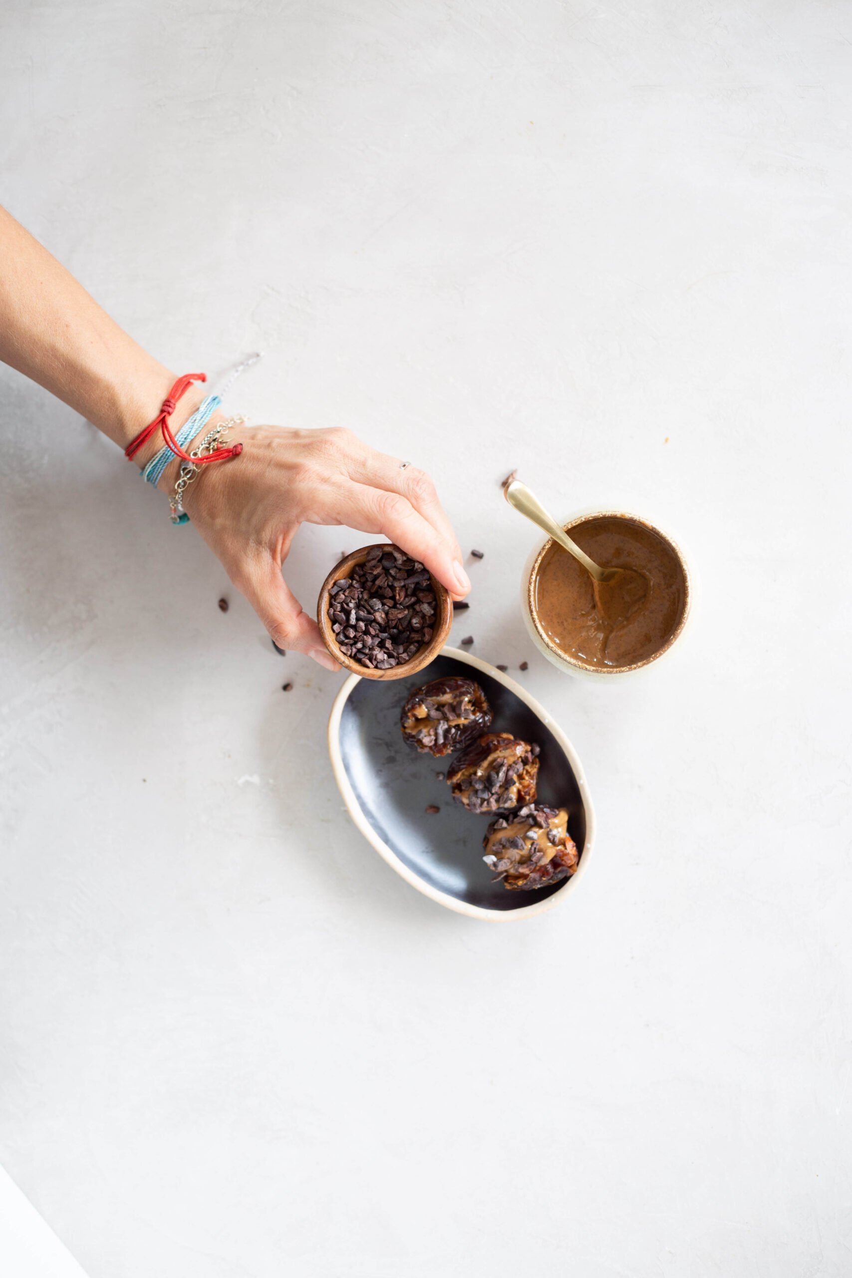 A hand wearing colorful bracelets holds a small bowl of cacao nibs above a white surface. Next to it are a cup of brown paste or sauce and an oval dish with three chocolate-covered almond butter stuffed dates. Some cacao nibs are scattered on the table.