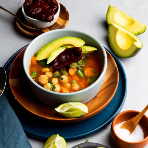 A bowl of comforting caldo tlalpeño garnished with avocado slices, served on a wooden plate. Nearby sits a small bowl of chipotle peppers, lime wedges, and a blue napkin. A dish of salt with a spoon and a plate of cilantro adorn the light-textured surface, completing this delicious Mexican soup setting.