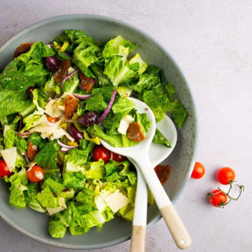 A green ceramic bowl filled with a fresh salad of lettuce, cherry tomatoes, sliced red onion, black olives, cheese slices, and dried fruit—perfect alongside your favorite Overnight Oats Recipe. Two white-handled servers rest in the bowl on a light countertop.