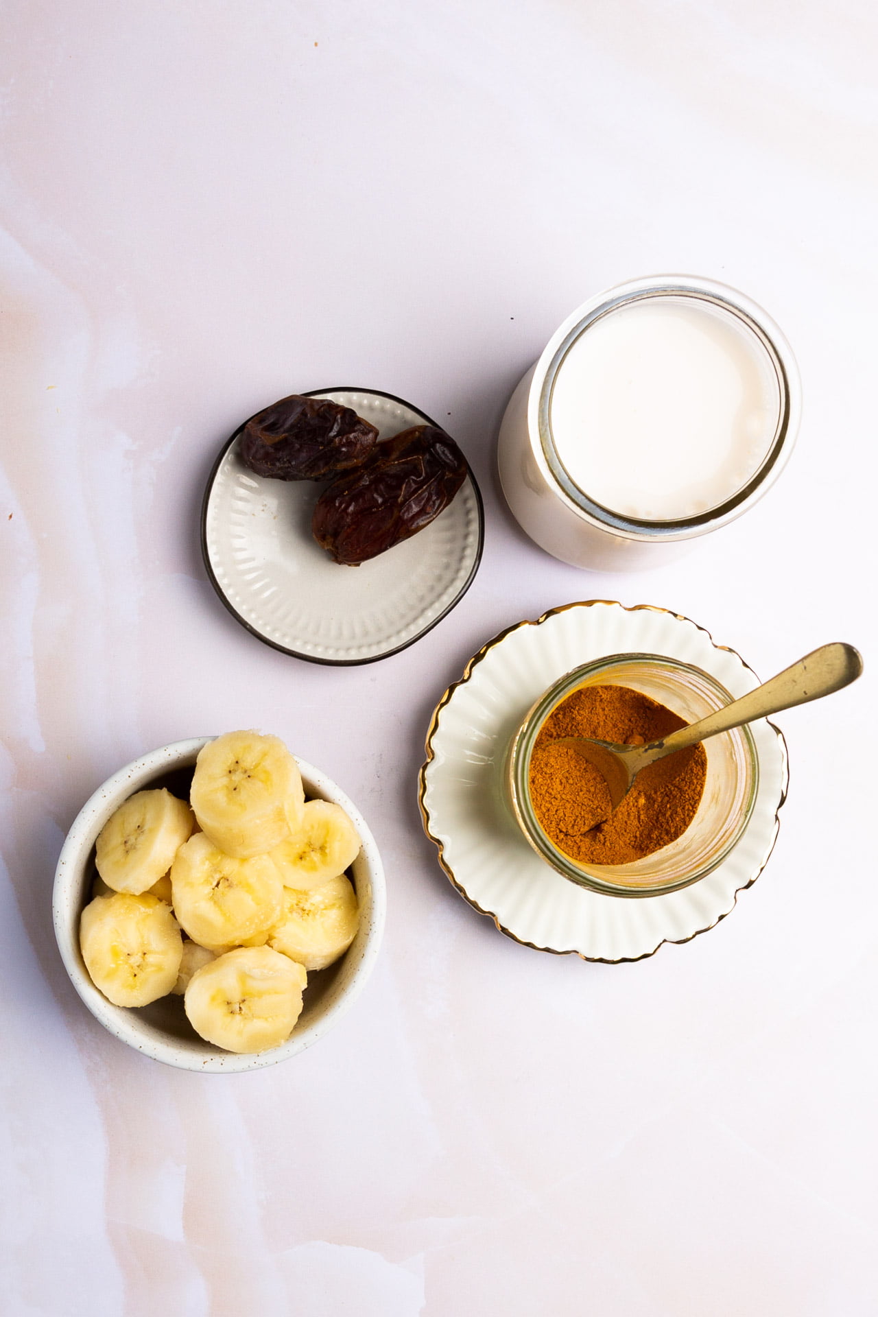 A flat lay of four ingredients for a Golden Banana Smoothie: sliced bananas in a bowl, two dates on a small plate, a glass of milk, and a jar of ground cinnamon with a spoon, all arranged on a light-colored surface.