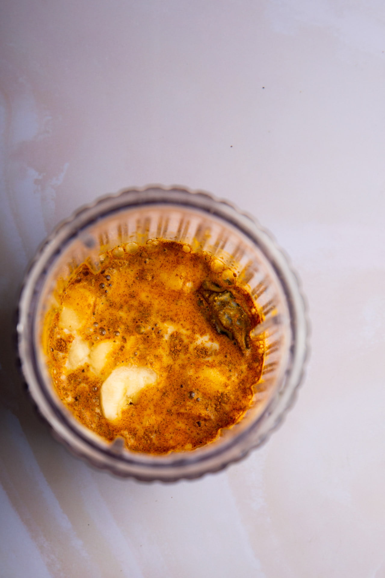 A top-down view of a clear glass filled with a vibrant yellow-orange liquid, resembling a Golden Banana Smoothie, with visible oil, seasonings, and chunks of white food—possibly cheese or tofu—on a light, marbled surface.