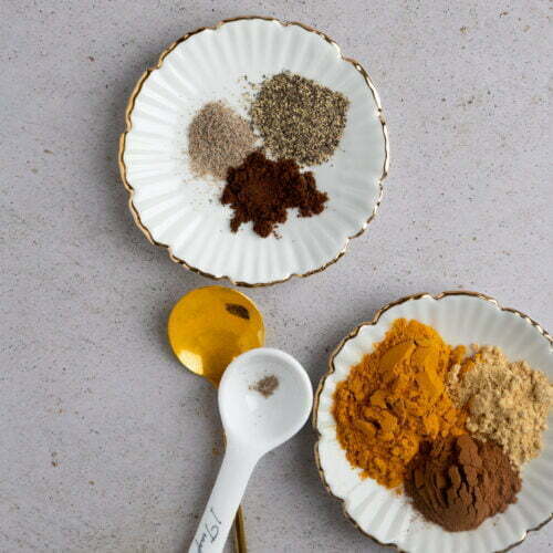 Two white scalloped dishes hold a colorful spice mix for golden milk, featuring ground pepper, cinnamon, turmeric, ginger, and nutmeg. A gold spoon and a white measuring spoon rest nearby on a light grey textured surface.