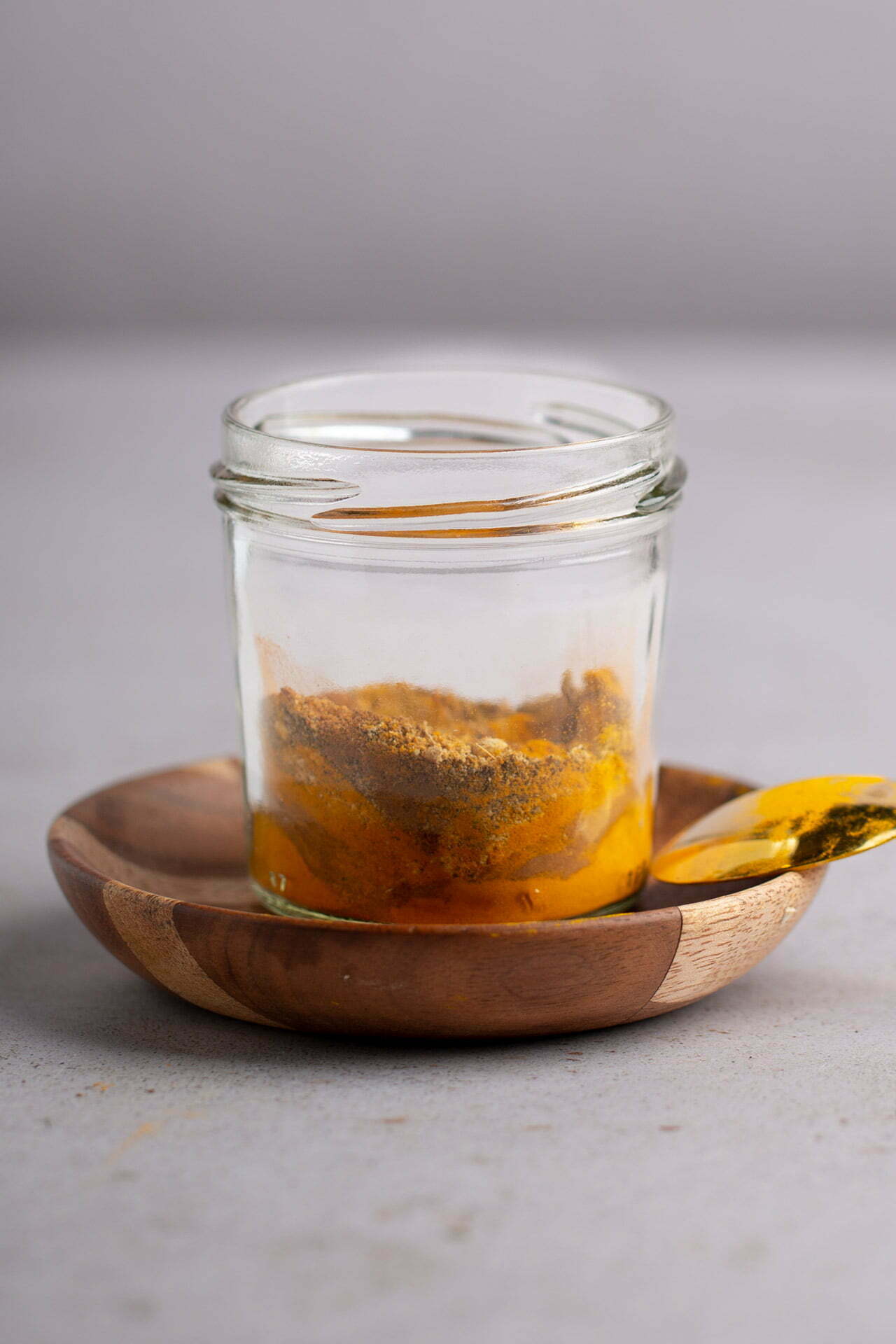 A small glass jar with yellow golden milk powder and brown spices sits on a round wooden dish. A spoon with the vibrant spice mix rests on the edge of the dish. The background is neutral and unfocused.
