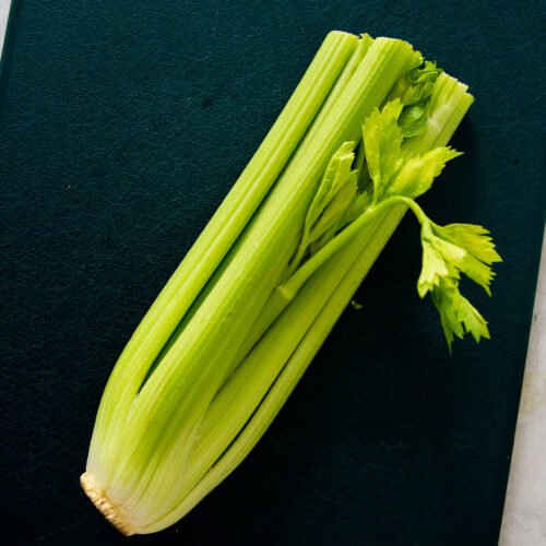 A bunch of fresh celery with leafy tops rests on a dark green cutting board, shown from above. Known for its health benefits, these light green stalks are uncut with the root end still attached. The board sits on a light countertop.