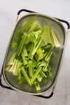 A metal colander filled with fresh celery stalks and leaves, cut into pieces, sits on a light gray countertop. The vibrant green celery, ideal for juicing to enjoy its benefits and properties, appears freshly washed with leafy tops visible.