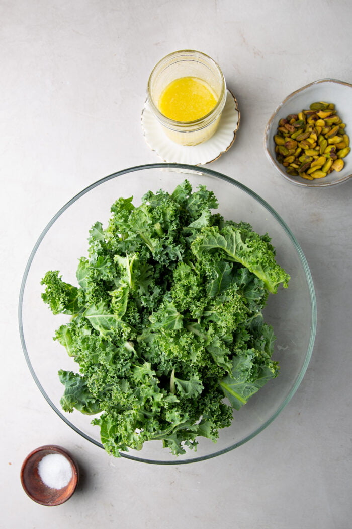 kale leaves in a salad bowl, dressing in a jar and pistachios in a bowl.