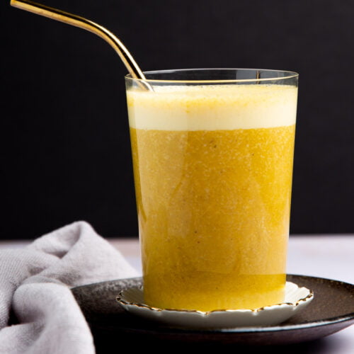 A glass of golden yellow turmeric latte with a frothy top sits on a black saucer, accompanied by a gold metal straw. A gray cloth is draped to the side, and a golden spoon with turmeric powder or Golden Banana Smoothie mix rests in the foreground.