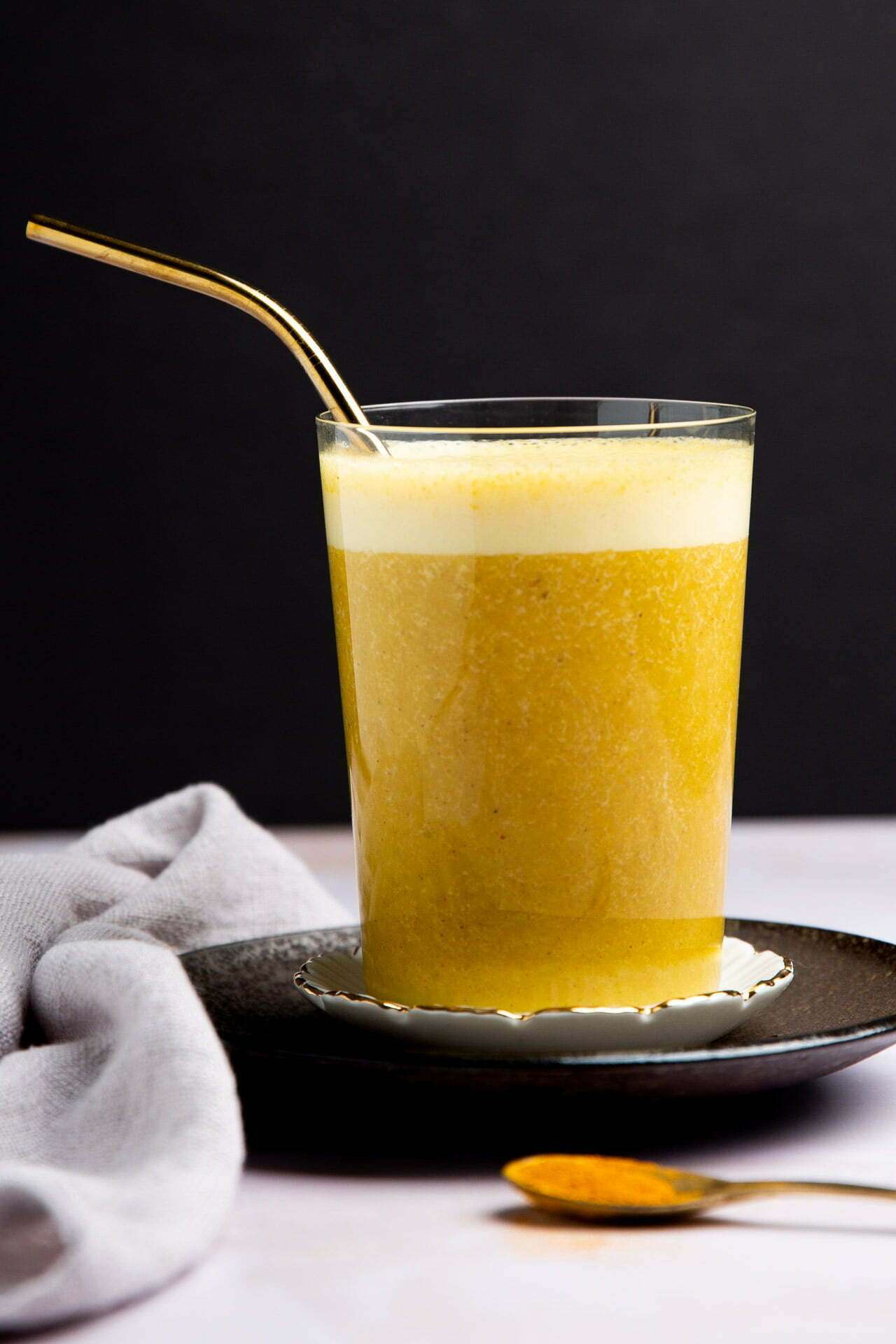 A glass of golden yellow turmeric latte with a frothy top sits on a black saucer, accompanied by a gold metal straw. A gray cloth is draped to the side, and a golden spoon with turmeric powder or Golden Banana Smoothie mix rests in the foreground.