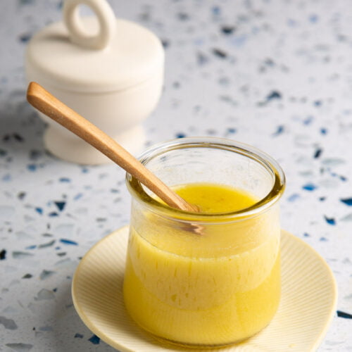 A glass jar filled with creamy, yellow ghee sits on a light-colored ceramic saucer. A wooden spoon rests inside the jar beside a tangy lemon dressing. In the background, there's a white, lidded container. The surface is a textured terrazzo with blue and gray speckles.