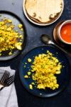 Two dark plates with perfectly scrambled tofu, herbs, and spices on a dark surface. Nearby are corn tortillas on a small plate, a bowl of red salsa, a spoon, and forks on a grey napkin. The scene is set for a meal conveying freshness and vibrant flavors.