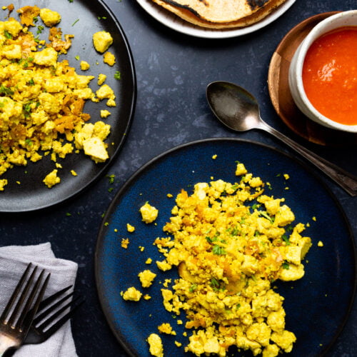 Plates of perfect scrambled tofu with herbs sit on a dark blue surface. A side dish of orange sauce accompanies them in a small bowl. Charred tortillas are stacked in the background. Nearby, a folded napkin with two forks and a spoon rests beside one plate.