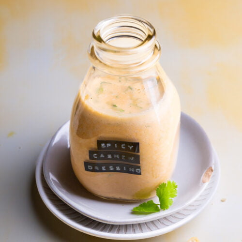 A small glass bottle filled with vibrant orange, Trader Joe's copycat spicy cashew dressing sits on stacked white plates. The bottle has a black label with white text. A cilantro leaf and some crushed cashew pieces are scattered around the plate on a light beige background.