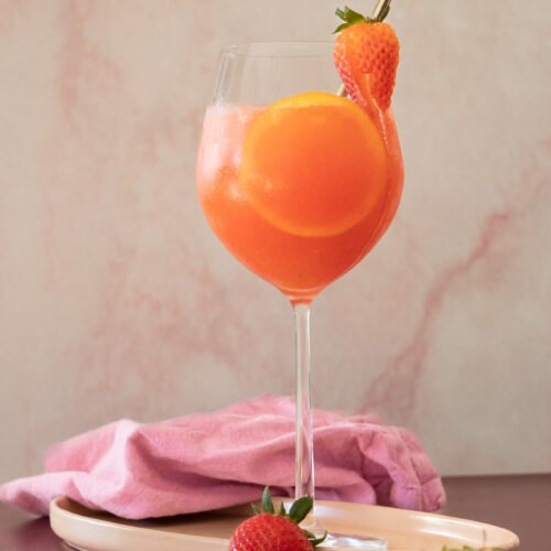 A wine glass filled with a vibrant orange-pink Strawberry Aperol Spritz, garnished with an orange slice and strawberry on the rim. A metal straw sits in the glass, while fresh strawberries and orange slices rest on a nearby plate.