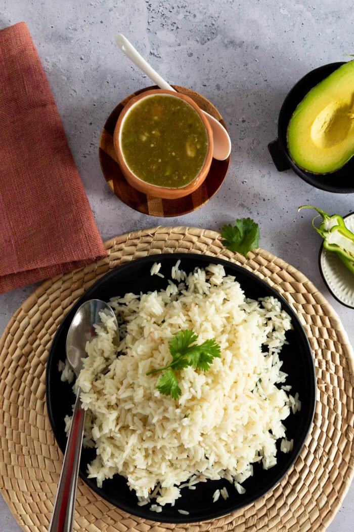 White rice on a black plate with salsa verde, tortillas and an avocado on the side