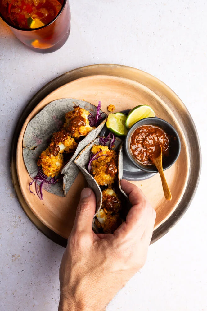 Una mano sostiene un taco de tortilla de ma&iacute;z azul relleno de pescado frito, col lombarda rallada y salsa. Otro taco, como los tacos de coliflor crujiente, reposa en un plato beige con salsa, una cuchara de madera, gajos de lima y una bebida c&iacute;trica cerca.