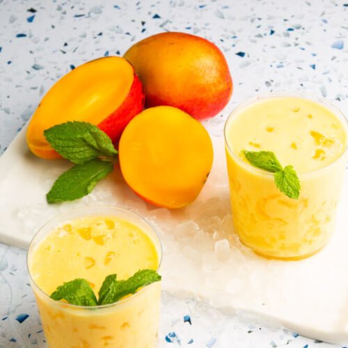 Two glasses of vegano mango lassi garnished with mint leaves rest on a white marble board with ice. Nearby, whole and halved ripe mangoes contrast beautifully against the speckled white surface, highlighting the vibrant orange hues of both the mangoes and lassi.