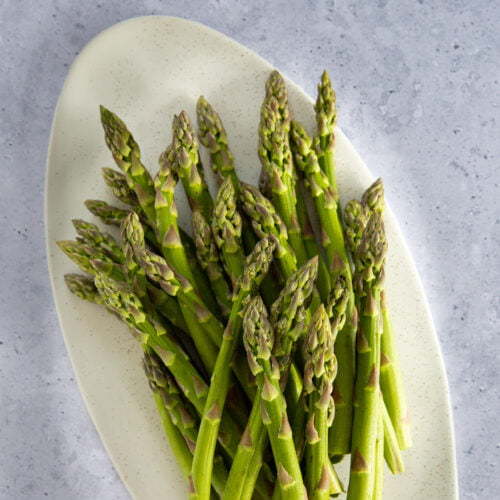 A bunch of sauteed asparagus spears is arranged neatly on an oval, off-white plate. The plate sits on a light gray, textured surface. The asparagus appears firm and vibrant, with tightly closed tips.