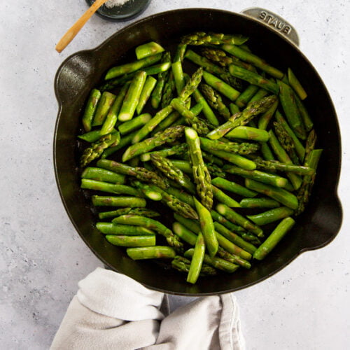 A black skillet filled with easy asparagus, sautéed to a bright green, sits on a light grey surface. A beige cloth is wrapped around one pan handle. Above the skillet, a small wooden bowl of coarse salt with a tiny spoon is placed.