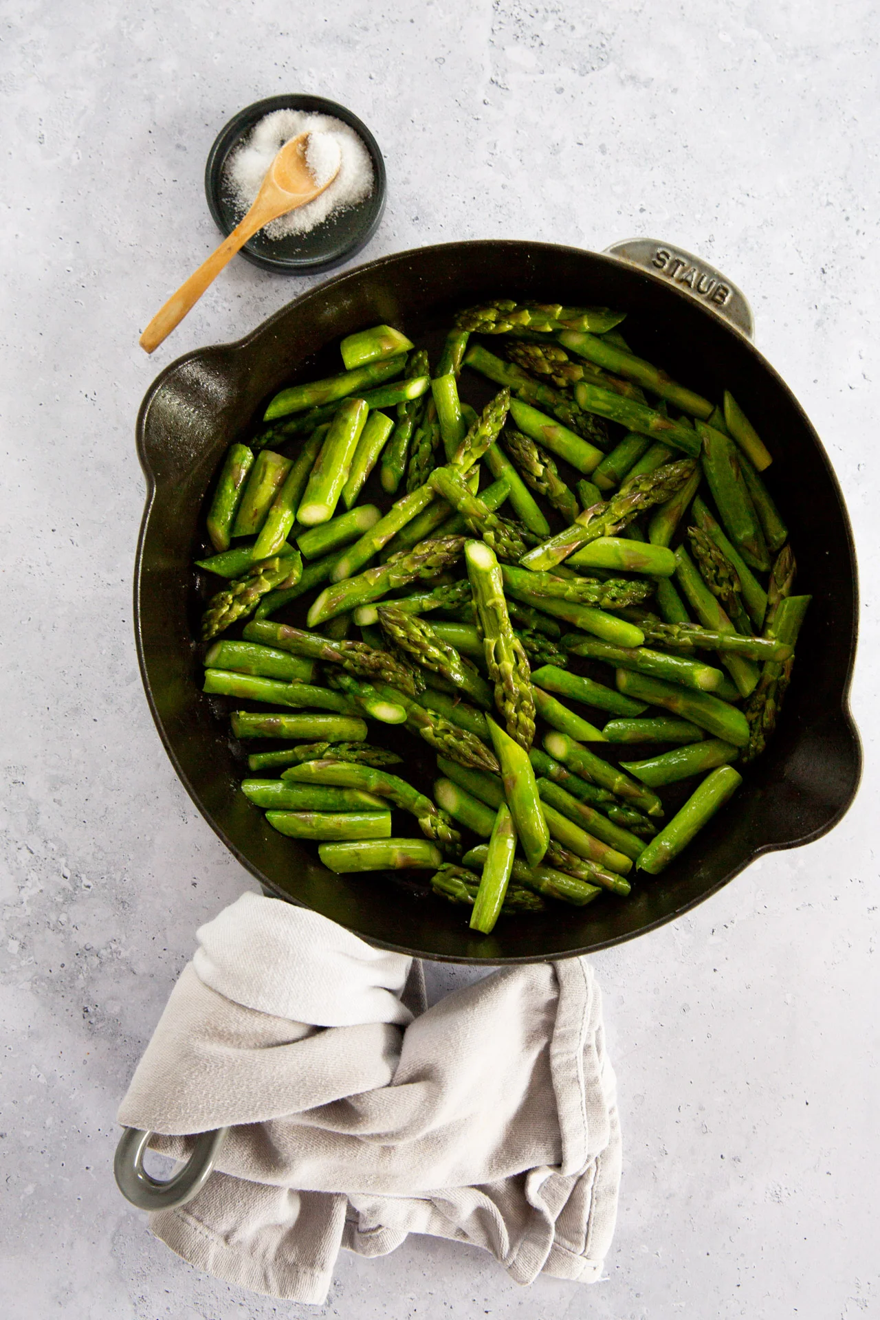A black skillet filled with easy asparagus, saut&eacute;ed to a bright green, sits on a light grey surface. A beige cloth is wrapped around one pan handle. Above the skillet, a small wooden bowl of coarse salt with a tiny spoon is placed.