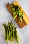 A bundle of fresh green asparagus spears, perfect for easy asparagus recipes like pan-fried asparagus, is arranged on a wooden cutting board with a leather loop, next to another bundle on an oval white plate atop a light gray speckled surface.