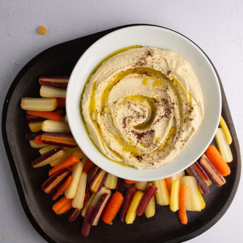 A bowl of basic hummus drizzled with olive oil and sprinkled with spices sits on a dark tray, surrounded by colorful carrot sticks in orange, yellow, and purple hues, arranged in a fan shape on a light gray background.