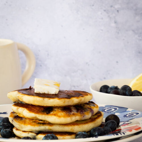 A plate with a stack of four Vegan Lemon Blueberry Pancakes topped with a small square of butter is surrounded by fresh blueberries. There's a mug on the left and a bowl of blueberries on the right, set against a light background.