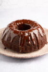 A luscious chocolate Bundt cake sits on a round, beige plate, dripping with glossy glaze that cascades down its sides. The subtle white and gray hexagonal pattern in the background accentuates the rich, dark chocolate hue of this decadent dessert.