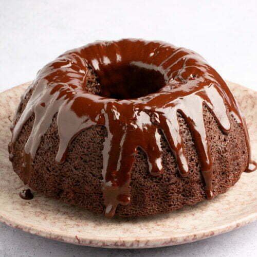 A luscious chocolate Bundt cake sits on a round, beige plate, dripping with glossy glaze that cascades down its sides. The subtle white and gray hexagonal pattern in the background accentuates the rich, dark chocolate hue of this decadent dessert.