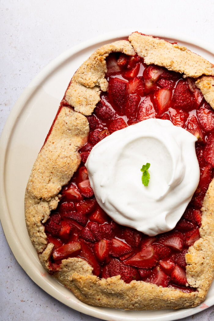 A rustic strawberry galette sits on a white plate, its golden-brown crust folded over juicy chopped strawberries. Reminiscent of the wholesome feel of a classic overnight oats recipe, a generous dollop of whipped cream with a mint garnish is on top, slightly off-center. The background is a light, textured surface.