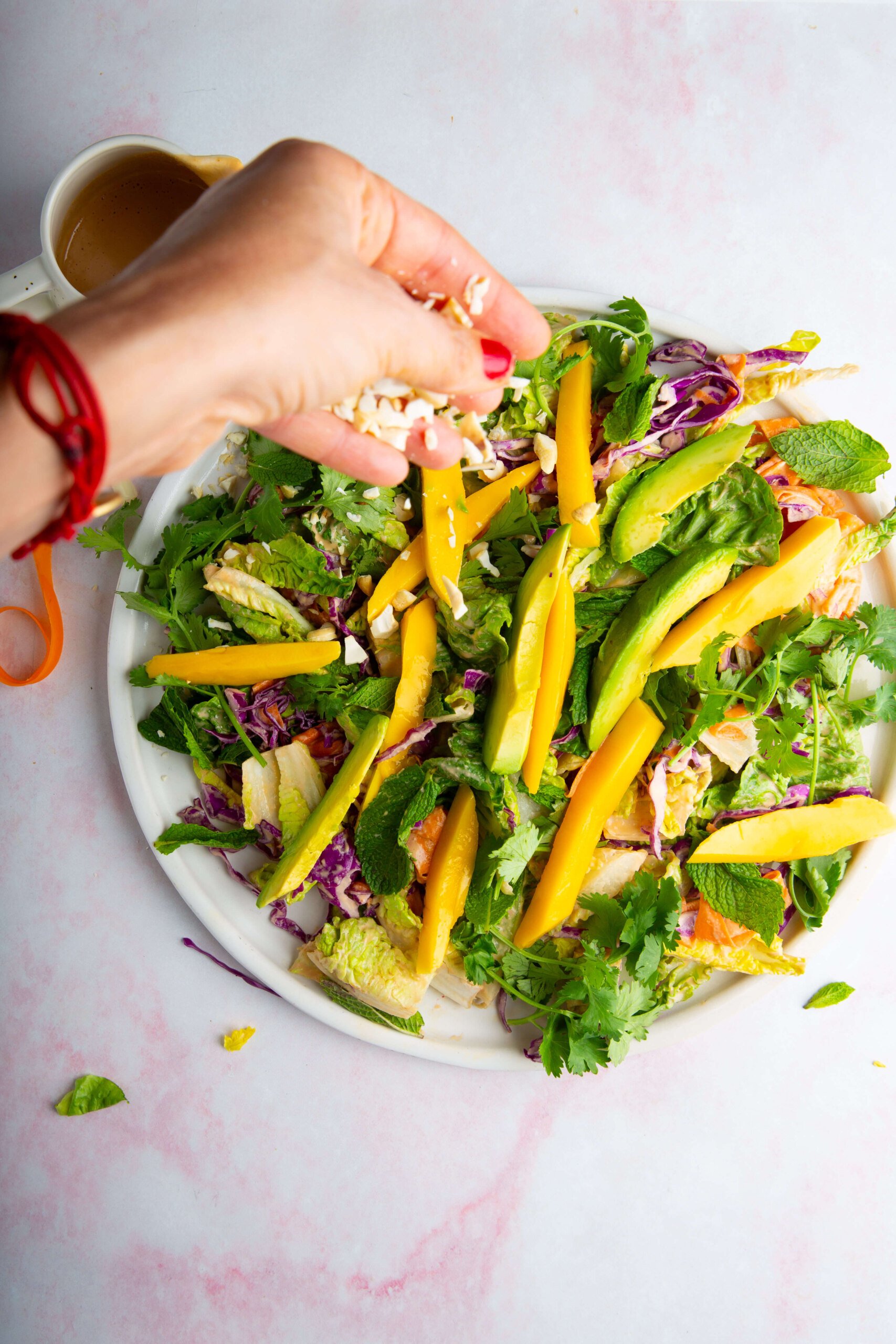 A hand with red nail polish sprinkles chopped nuts onto a colorful salad of greens, sliced mango, avocado, purple cabbage, and herbs on a white plate. A cup of dressing sits in the background on a light, pink-tinged surface.