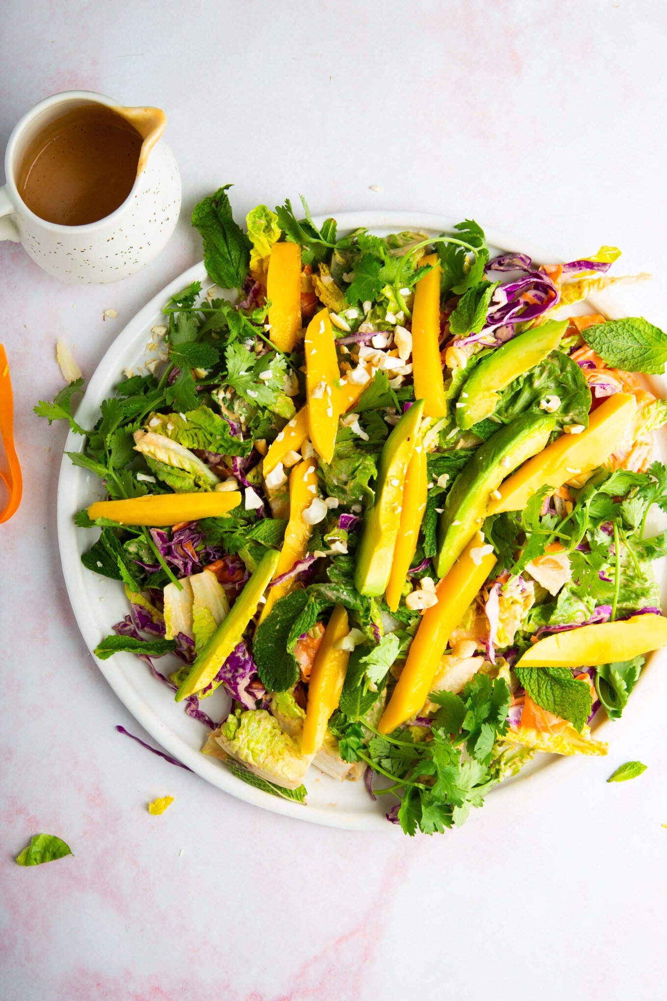 A vibrant ensalada crujiente on a white plate features sliced mango, avocado, shredded purple cabbage, greens, cilantro, and nuts. A small pitcher of aderezo de cashew sits nearby on a light pink and white surface. The salad looks fresh and colorful.