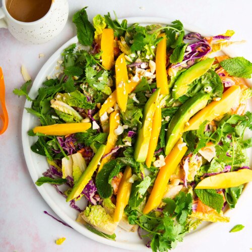 A vibrant ensalada crujiente on a white plate features sliced mango, avocado, shredded purple cabbage, greens, cilantro, and nuts. A small pitcher of aderezo de cashew sits nearby on a light pink and white surface. The salad looks fresh and colorful.