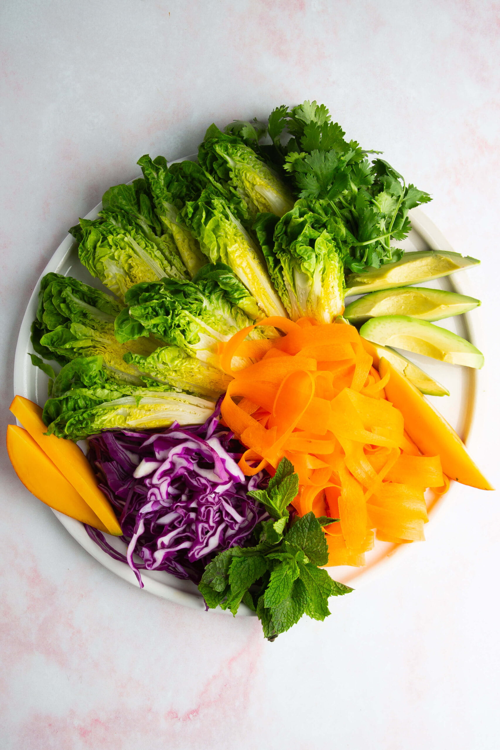 A white plate holds arranged fresh vegetables and fruits: baby romaine lettuce, cilantro, avocado slices, carrot ribbons, purple cabbage shreds, mango slices, and mint sprigs. The background is a light pinkish-white marble surface.