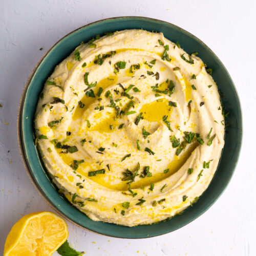 A bowl of creamy Lemon Hummus with mint, drizzled with olive oil and garnished with chopped herbs, sits on a light surface. Beside the bowl, a halved lemon and a fresh sprig of mint highlight its smooth texture and swirl pattern.