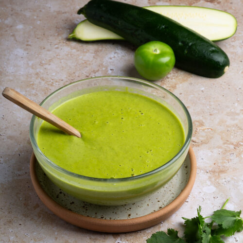A glass bowl of green soup with a wooden spoon sits on a beige plate. Behind it are two zucchinis, one sliced, and a green tomatillo—perfect ingredients for Calabacita salsa recipe inspiration. Fresh cilantro leaves rest in the foreground.